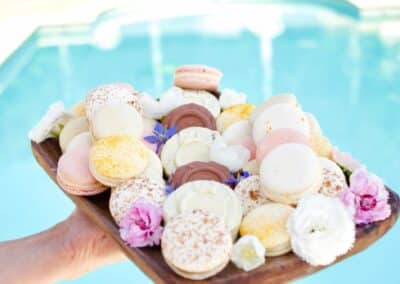 Une personne tient un plateau de macarons colorés et fleurs, avec une piscine en arrière-plan, évoquant une ambiance estivale et festive.