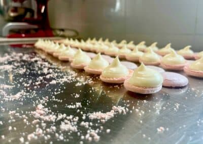 Des coques de macarons alignées sur une plaque en métal, garnies d'une crème. Des miettes roses parsèment la surface brillante.