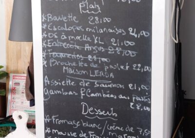 Un menu du jour écrit à la craie sur un tableau noir dans un café, avec une lampe et des éléments de décor.