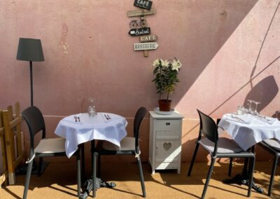 Deux tables avec nappes blanches dans un café en plein air, décor rustique avec signes et plantes, mur rose en arrière-plan.