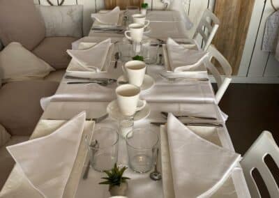 Table élégamment dressée avec nappes blanches, tasses et assiettes. Décoration chaleureuse et rustique, ambiance conviviale et accueillante, prête pour des invités.