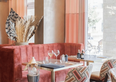 Un coin de restaurant élégant avec une banquette rouge, décoration de plumes, trois verres à pied et des chaises colorées près d'une fenêtre ensoleillée.