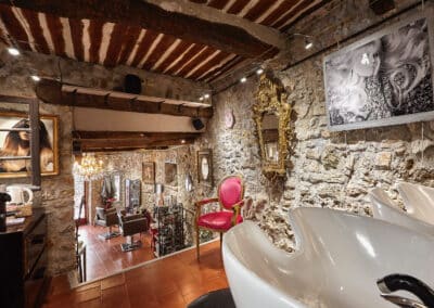 Un salon de coiffure élégant avec murs en pierre, décoration vintage, miroirs dorés, fauteuil rose, œuvres d'art et luminaires suspendus. Ambiance chaleureuse.