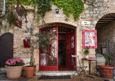 Un café pittoresque en pierre avec une entrée rouge, des plantes en pot et une affiche encadrée d'une personne, situé rue des Lombards.