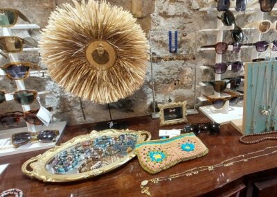 Accessoires exposés sur un comptoir en bois: lunettes de soleil, bijoux, pochette en crochet et décoration murale en paille sur fond de pierre.
