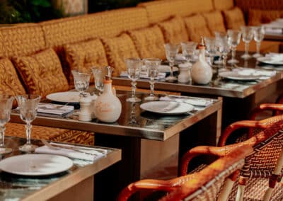 Un restaurant élégant avec des tables bien dressées, des verres en cristal, des assiettes blanches et des sièges en rotin, créant une atmosphère accueillante.