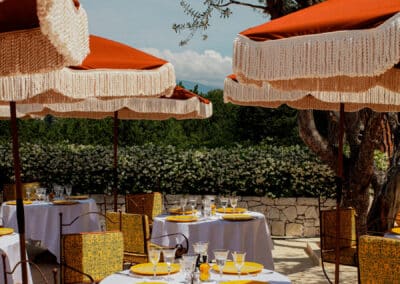 Tables élégamment dressées sous parasols rouges dans un restaurant en terrasse, entourées de verdure. Atmosphère chic et décontractée, idéale pour un repas.