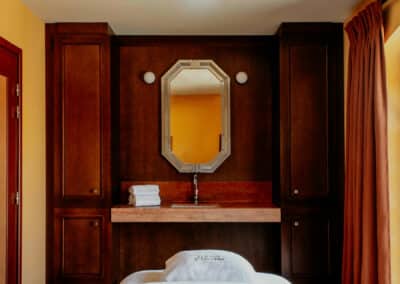 Une salle de spa élégante avec table de massage, serviettes blanches, décor bois, ventilateur au plafond, miroir octogonal, et rideaux orange.