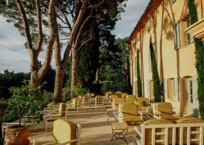 Terrasse tranquille avec sièges ensoleillés, grands arbres et façade historique provençale. L'atmosphère méditerranéenne invite à la détente en plein air.