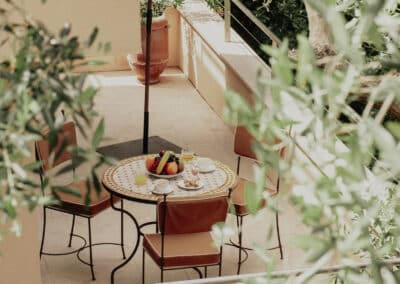 Un balcon ensoleillé avec table, chaises et parasol rouge, entouré de verdure. Petit déjeuner avec fruits et jus sur la table.