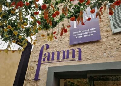 Un bâtiment en pierre avec une enseigne "famm". Des fleurs suspendues décorent l'entrée. Premier musée privé dédié aux artistes femmes en Europe.