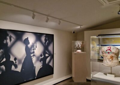 Une exposition d'art avec des silhouettes de personnes, un buste décoré et des objets lumineux sous verre, dans une galerie moderne et minimaliste.