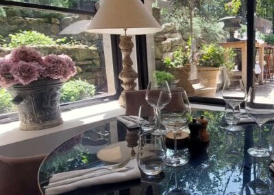 Une table élégante avec des verres à vin, lampe et fleurs roses. Vue extérieure verdoyante, ambiance lumineuse et agréable dans un cadre paisible.