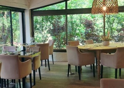 Salle de restaurant lumineuse avec grandes fenêtres donnant sur jardin verdoyant, meubles en bois, chaises confortables et suspension en osier au plafond. Ambiance paisible.