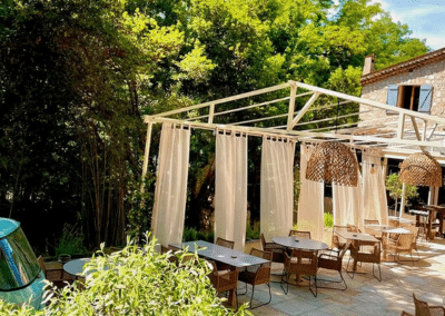 Un restaurant extérieur charmant avec des rideaux blancs, des tables en osier, entouré de verdure et d'un bâtiment en pierre, sous un ciel ensoleillé.