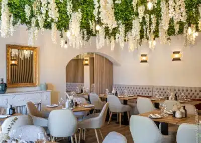 L'intérieur d'un restaurant élégant avec tables en bois, plafond décoré de fleurs blanches et verdure, ambiance chaleureuse et moderne, sans personne visible.