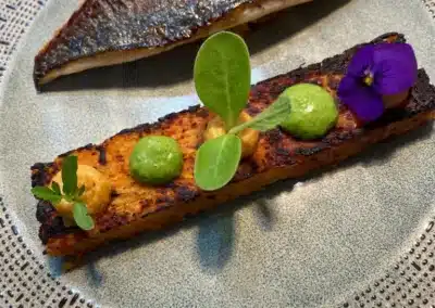 Un plat élégant avec poisson grillé et garniture, décoré de sauce verte, fleur comestible, sur une assiette en céramique texturée.