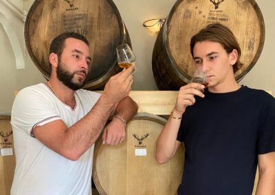 Two people taste whisky in front of wooden barrels, concentrating on the aroma in their glass, in a sober, warm setting.