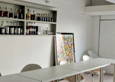 Salle lumineuse avec étagères remplies de bouteilles, tableau coloré et bureaux blancs. Fenêtre avec vue extérieure. Ambiance moderne et épurée.