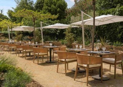 Terrasse de restaurant extérieure avec chaises en osier et tables dressées, entourée de verdure luxuriante. Parapluies blancs offrent de l'ombre. Ambiance paisible.