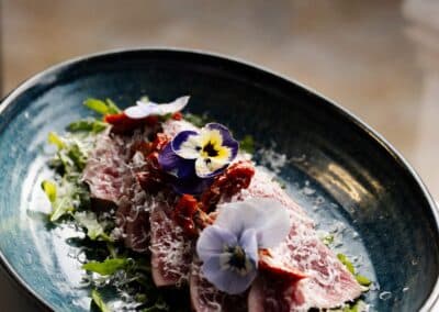 Un plat élégant avec du rôti garni de fleurs comestibles et de roquette, présenté dans une assiette bleue sur une table lumineuse.