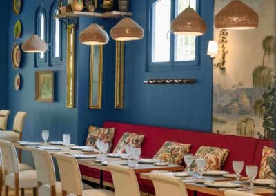 Un restaurant coloré avec murs bleus, décorations murales, table dressée, coussins floraux, ambiance conviviale, éclairage suspendu, et sièges confortables.