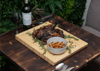 Une table en bois avec poulet rôti, frites, herbes, bouteille de vin rouge et verre, située devant un mur végétal.