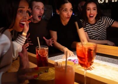 Quatre personnes autour d'une table de jeu, expressions surprises, boissons colorées devant eux. En arrière-plan, le logo "UMAI" est illuminé.