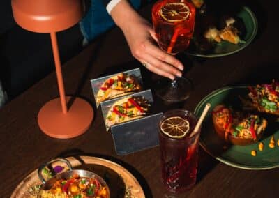 Une personne savoure des cocktails colorés avec des tacos et plats variés, éclairée par une lampe, sur une table en bois sombre.