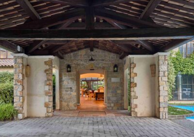 Un porche en pierre mène à un intérieur cozy avec fauteuils. Végétation extérieure, piscine partiellement visible à droite. Atmosphère accueillante et rustique.