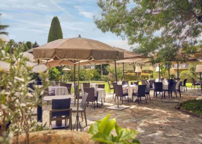 Terrasse de restaurant élégante avec tables dressées, parasols ombragés et jardin verdoyant en toile de fond, par une journée ensoleillée. Atmosphère paisible et accueillante.