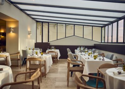 Un restaurant élégant avec des tables dressées, sous une véranda en verre. Ambiance lumineuse et moderne, chaises en bois et verres de vin.