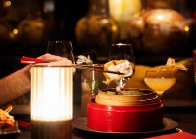 Personne dégustant un plat dans un cadre élégant avec vase décoratif et lumière tamisée, ambiance asiatique mise en valeur par les baguettes rouges.