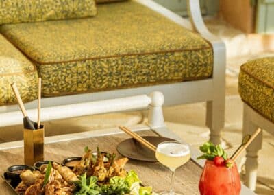 Plateau de mets variés avec deux cocktails colorés sur une table en bois, à côté d'un canapé élégant à motifs jaunes. Ambiance estivale.