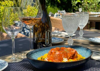 Dîner en plein air avec verre de vin, bouteille de Château et plat coloré sur table décorée, entouré de nature verdoyante.
