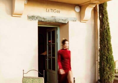 Une personne en robe rouge sort de "Le Tigrr", terrasse avec chaises et table en métal, décor méditerranéen chaleureux.