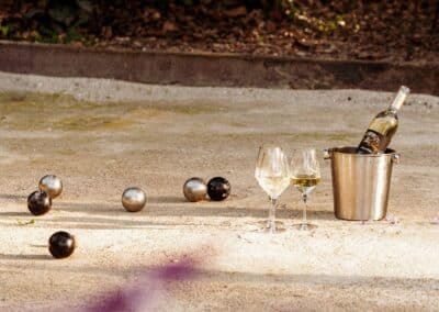 Bouteille de vin, verres, et boules de pétanque sur un terrain sablé, entourés de végétation et lumière douce. Ambiance décontractée et sereine.