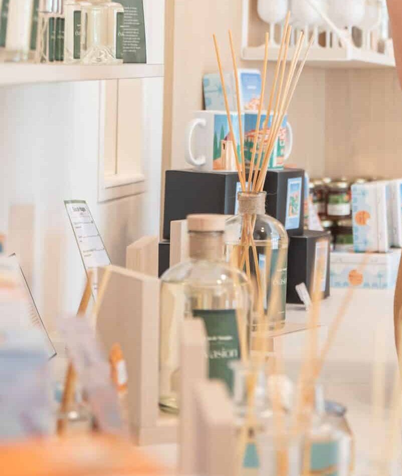 Une personne dans une boutique sent un produit, entourée de diverses marchandises exposées sur des étagères. Ambiance chaleureuse et lumineuse.