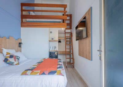 Chambre d'hôtel moderne avec mezzanine en bois, lit double confortable, oreillers colorés, télévision murale, et échelle menant à un lit supplémentaire en hauteur.