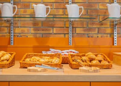 Cinq paniers de viennoiseries variées sur une table en bois, devant un mur en briques. Des pichets blancs sont posés sur une étagère.