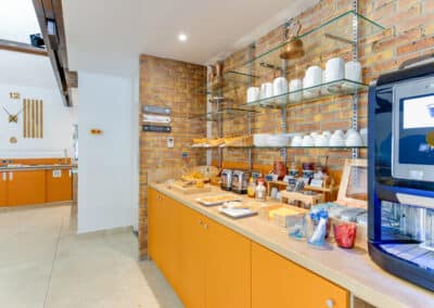 Un coin cuisine moderne avec des étagères en verre, des tasses blanches, et un distributeur de boissons. Murs en briques et meubles orange.