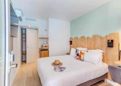 Chambre lumineuse avec lit double, décoration moderne, chapeau et magazines sur le lit, mur bleu clair, et meuble avec bouilloire dans le coin.