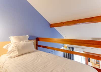 Un lit mezzanine avec draps blancs, situé sous un plafond incliné bleu. L'éclairage doux crée une ambiance chaleureuse et accueillante.