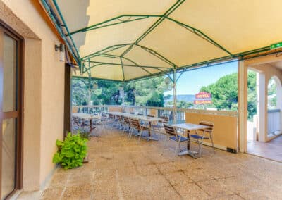 Terrasse extérieure d'un hôtel avec tables et chaises sous une pergola. En arrière-plan, panneaux "Hôtel Belvédère" et arbres verdoyants.