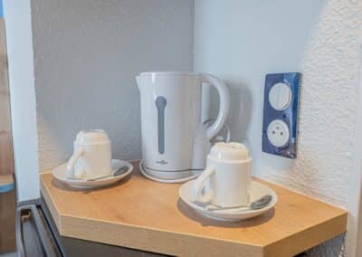 Une bouilloire blanche sur l'étagère en bois avec deux tasses blanches, posées près d'une prise électrique murale.