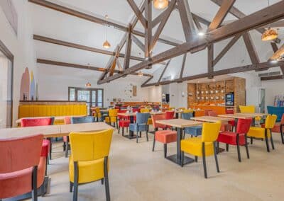Une salle à manger colorée avec des tables en bois, des chaises jaunes, rouges et bleues, et de grandes poutres en bois apparentes.