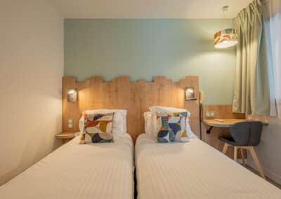 Chambre moderne avec deux lits simples, décorée de coussins géométriques colorés, une lampe suspendue, bureau en bois et chaises élégantes. Atmosphère chaleureuse et accueillante.