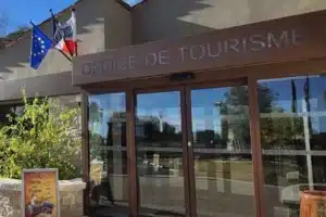Entrée moderne de l'office de tourisme, entourée de verdure et murs en pierre, sous un ciel partiellement nuageux.